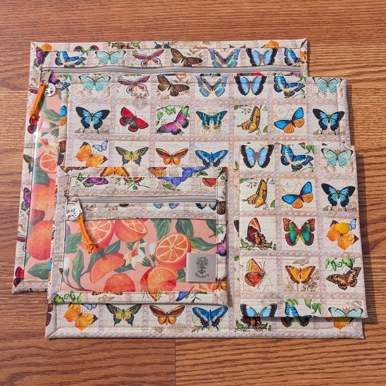 May include: A set of three zippered pouches with a butterfly and orange slice pattern. The largest pouch features a grid of colorful butterflies. The middle pouch has a clear window with orange slices. The smallest pouch has a butterfly pattern.