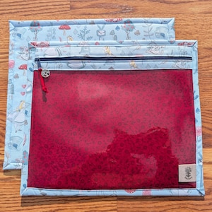 Könnte beinhalten: Zwei Reißverschlusstaschen. Die obere Tasche hat einen hellblauen Stoff mit einem Alice im Wunderland-Thema. Die untere Tasche hat eine transparente rote Vorderseite. Beide Taschen haben einen dunkelblauen Reißverschluss und ein kleines Etikett.