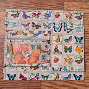 May include: A beige quilted organizer with a butterfly postage stamp design. It features a zippered pocket with an orange slice print and a small tag that reads "Candy Coffee Stickers". The organizer is on a wooden surface.