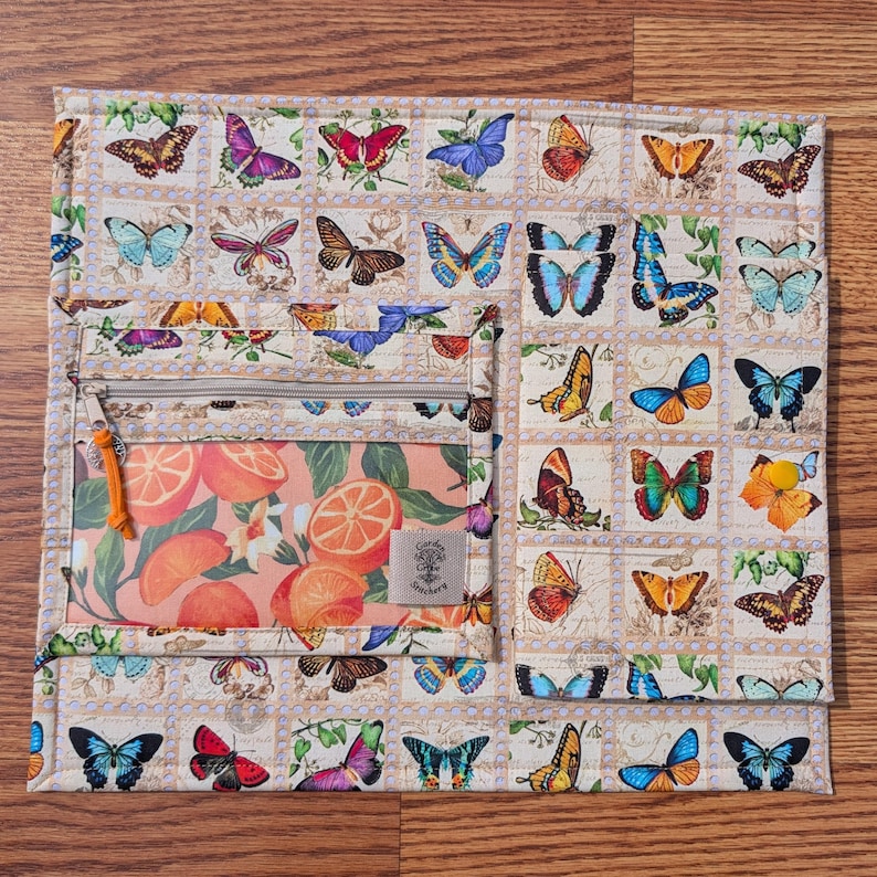 May include: A beige quilted organizer with a butterfly postage stamp design. It features a zippered pocket with an orange slice print and a small tag that reads "Candy Coffee Stickers". The organizer is on a wooden surface.