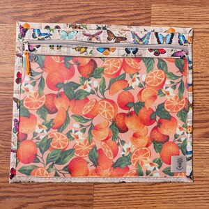 May include: A rectangular zippered pouch with a vibrant orange and green citrus fruit pattern. The top section features a butterfly print. The pouch has a beige zipper and an orange pull. The corners are reinforced with a patterned fabric.