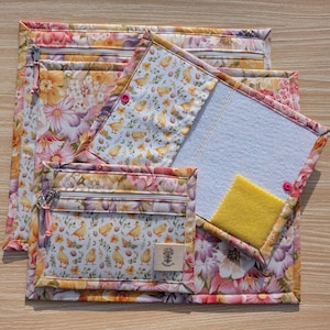 Puede incluir: Un conjunto de bolsas de tela acolchada y una funda para bloc de notas. El estampado floral presenta flores rosas, amarillas y moradas. Una bolsa tiene una ventana transparente y una cremallera. La funda del bloc de notas tiene un cuadrado de fieltro amarillo y un cierre de bot&oacute;n.