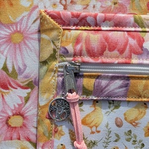 Puede incluir: Primer plano de una tela floral con cremallera y un charm. La tela presenta flores rosas y amarillas, con una cremallera blanca y un charm de &aacute;rbol de la vida plateado. Un cord&oacute;n rosa est&aacute; unido a la cremallera.