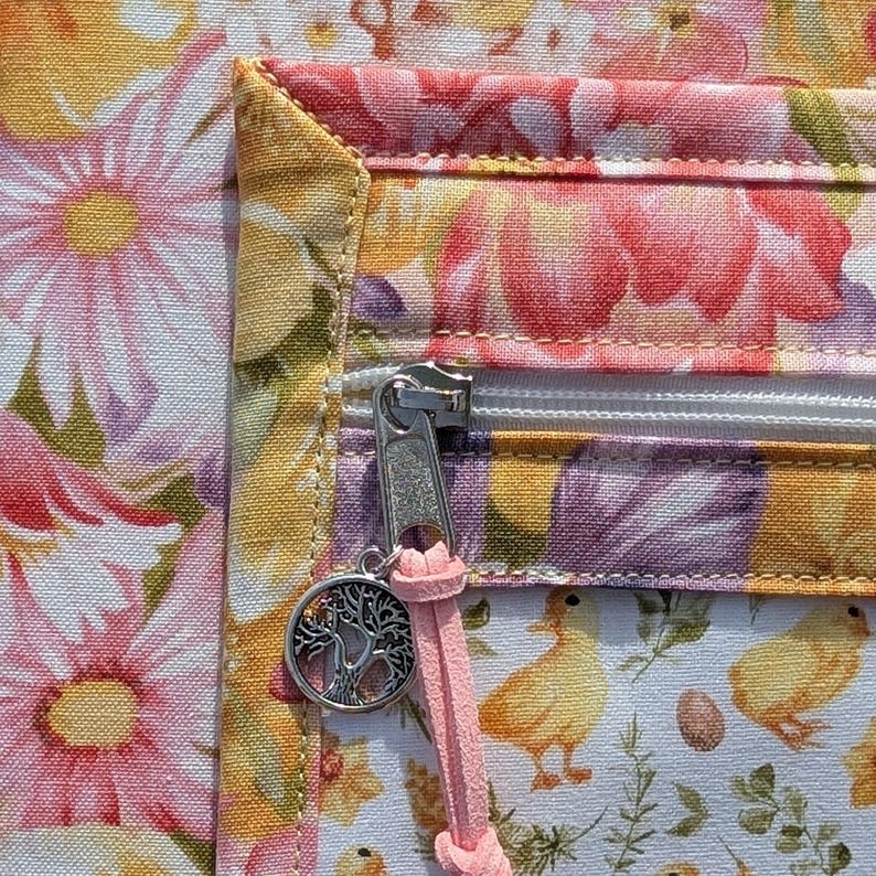 Puede incluir: Primer plano de una tela floral con cremallera y un charm. La tela presenta flores rosas y amarillas, con una cremallera blanca y un charm de &aacute;rbol de la vida plateado. Un cord&oacute;n rosa est&aacute; unido a la cremallera.