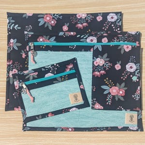 May include: A set of four fabric pouches with a floral pattern on a black background. The pouches have teal zippers and a teal interior. The floral design includes pink and white flowers with green leaves. Each pouch has a small tag.