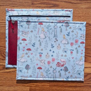 May include: Two fabric pouches with an Alice in Wonderland theme. The larger pouch has a light blue background with illustrations of Alice, mushrooms, and clocks. The smaller pouch has a red panel with a zipper and a charm.