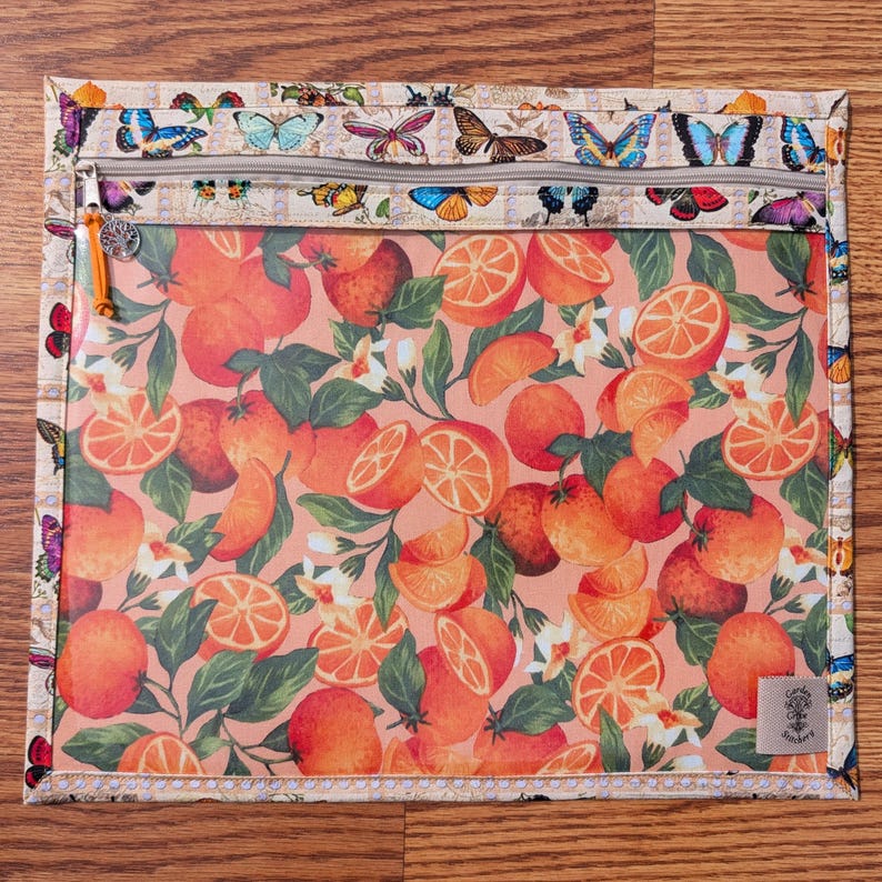 May include: A square, zippered pouch featuring a vibrant orange and green citrus fruit pattern on a peach background. The top border has a butterfly design. The pouch has a zipper with an orange pull and a small tag that says "Garden Stitchery".