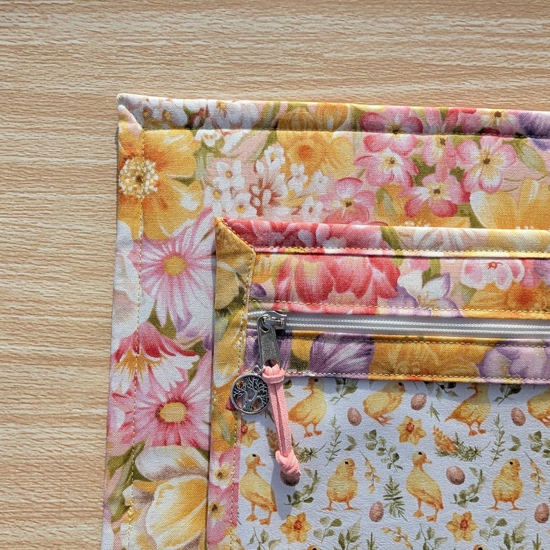 Puede incluir: Una bolsa de tela floral con cierre de cremallera. La bolsa presenta un patr&oacute;n vibrante de flores rosas, amarillas y blancas. El forro interior muestra un patr&oacute;n de patitos y huevos amarillos. Un cord&oacute;n rosa con un dije est&aacute; unido a la cremallera.