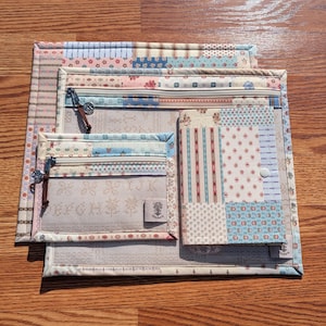 May include: A set of three fabric pouches with patchwork patterns in pastel colors. Each pouch has a zipper closure and decorative pulls. The smallest pouch has the letters "EFGH" visible. The pouches are arranged on a wooden surface.