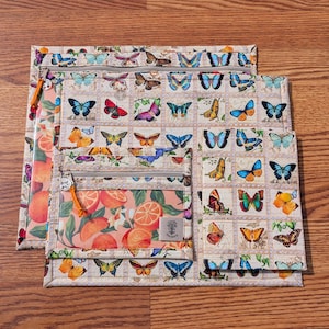 May include: Three zippered pouches with a butterfly and citrus fruit design. The largest pouch features a repeating pattern of colorful butterflies on a cream background. The smaller pouches have a citrus fruit pattern and a clear pocket. The zippers have orange pulls.