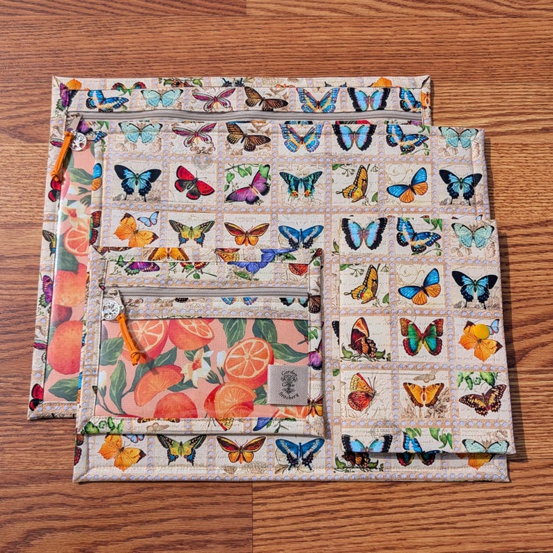 May include: Three zippered pouches with a butterfly and citrus fruit design. The largest pouch features a repeating pattern of colorful butterflies on a cream background. The smaller pouches have a citrus fruit pattern and a clear pocket. The zippers have orange pulls.