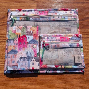 Könnte beinhalten: Ein Set aus drei Stoffbeuteln mit buntem Dorfaufdruck. Die Beutel haben transparente Vinyltaschen, Reißverschlüsse und einen Knopfverschluss. Der größte Beutel misst ca. 30 x 23 cm, der mittlere 23 x 18 cm und der kleinste 18 x 13 cm.