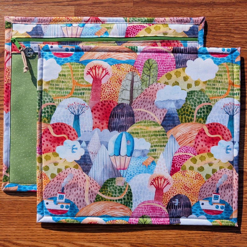 Puede incluir: Dos bolsas de tela coloridas con un dise&ntilde;o de paisaje. La bolsa m&aacute;s grande tiene una cremallera y un interior verde. El paisaje presenta monta&ntilde;as, &aacute;rboles, barcos y un globo aerost&aacute;tico.