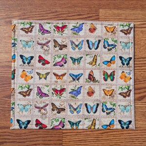 May include: A square, quilted placemat featuring a repeating pattern of colorful butterflies in various designs. The butterflies are set against a cream-colored background with a border of small squares. The placemat is on a wooden surface.