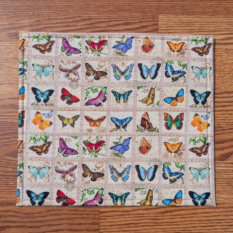 May include: A square, quilted placemat featuring a repeating pattern of colorful butterflies in various designs. The butterflies are set against a cream-colored background with a border of small squares. The placemat is on a wooden surface.