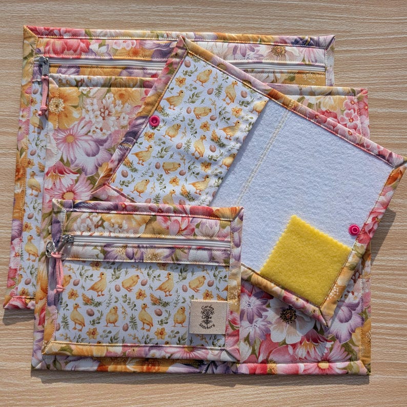 Puede incluir: Un conjunto de bolsas de tela acolchada y una funda para bloc de notas. El estampado floral presenta flores rosas, amarillas y moradas. Una bolsa tiene un frente transparente y una cremallera. La funda del bloc de notas tiene un cuadrado amarillo y un interior blanco.