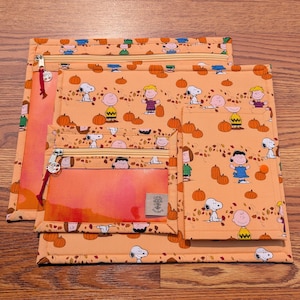 May include: A set of three orange fabric pouches featuring a Peanuts character print with Snoopy, Charlie Brown, and Lucy among pumpkins and autumn leaves. The pouches include zippered closures with silver charms and a clear vinyl window section.
