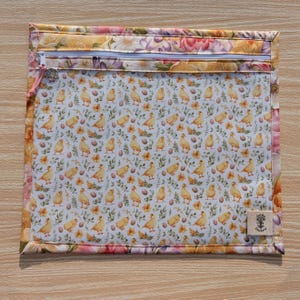 Puede incluir: Una bolsa rectangular con cremallera, borde floral e interior blanco estampado con pollitos amarillos, huevos y flores. La cremallera es blanca con un tirador rosa. Una peque&ntilde;a etiqueta con texto est&aacute; en la esquina inferior derecha.