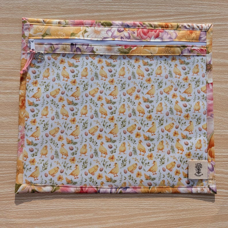 Puede incluir: Una bolsa rectangular con cremallera, borde floral e interior blanco estampado con pollitos amarillos, huevos y flores. La cremallera es blanca con un tirador rosa. Una peque&ntilde;a etiqueta con texto est&aacute; en la esquina inferior derecha.