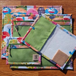 Puede incluir: Un conjunto de bolsas de tela coloridas con un patr&oacute;n de paisaje caprichoso. Las bolsas presentan interiores verdes con lunares, bolsillos con cremallera y un cierre de bot&oacute;n. La bolsa abierta revela un interior blanco y un peque&ntilde;o cuadrado de fieltro marr&oacute;n.