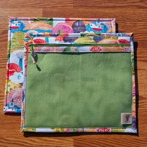 Puede incluir: Dos bolsas de tela con un colorido estampado de paisaje. La bolsa superior tiene una cremallera verde y un frente transparente. La bolsa inferior es visible, con un patr&oacute;n de paisaje similar. El patr&oacute;n incluye barcos, &aacute;rboles y formas coloridas.