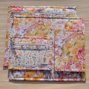 Puede incluir: Un conjunto de bolsas de tela acolchada con un estampado floral en tonos rosas, amarillos y naranjas. Las bolsas son de diferentes tama&ntilde;os y tienen cierres de cremallera y un peque&ntilde;o bolsillo con un estampado de pollitos y huevos. Una peque&ntilde;a etiqueta es visible.