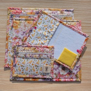 Puede incluir: Un conjunto de bolsas de tela con estampado floral y un bloc de notas. Las bolsas tienen cremalleras y un dise&ntilde;o de pollitos y huevos amarillos sobre fondo blanco. El bloc de notas tiene un interior blanco y un cuadrado amarillo.