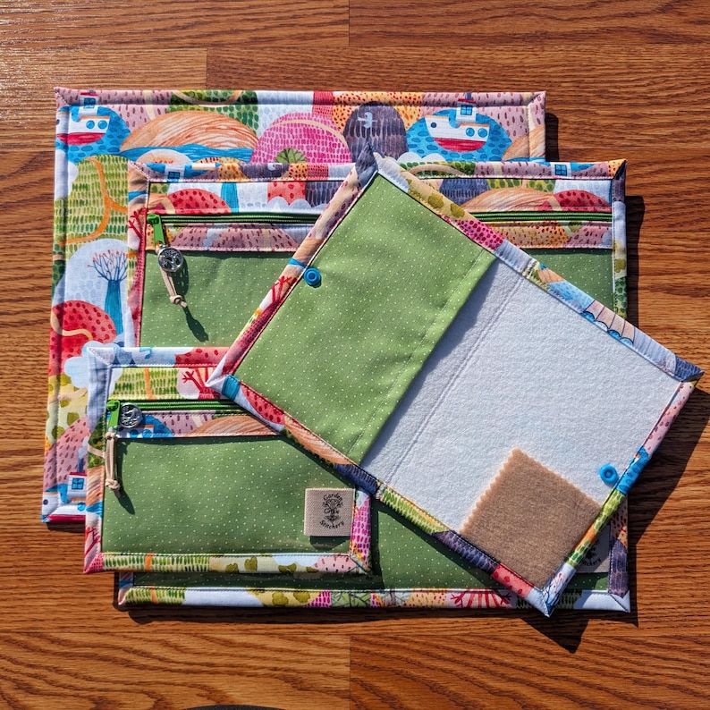 Puede incluir: Un conjunto de bolsas de tela coloridas con un patr&oacute;n de paisaje caprichoso. Las bolsas tienen bolsillos interiores verdes con cremalleras y cierres de bot&oacute;n azules. Una bolsa est&aacute; abierta, revelando un interior blanco y un peque&ntilde;o cuadrado de fieltro marr&oacute;n.