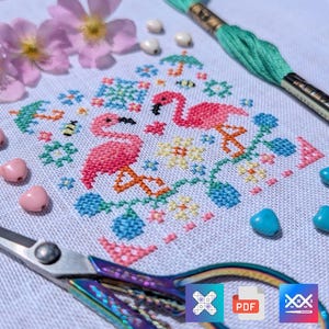 Peut inclure: Un projet de broderie au point de croix sur tissu blanc représentant deux flamants roses, des petites fleurs et des abeilles. Le motif est entouré de perles roses et bleues, d'un écheveau de fil turquoise et de ciseaux en métal irisé.