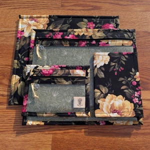 Puede incluir: Un conjunto de bolsas de tela acolchada y un folio. Los artículos presentan un estampado floral negro con flores amarillas y rosas. Las bolsas tienen bolsillos de vinilo transparente con cremalleras y una pequeña etiqueta. El folio tiene un cierre de botón rosa.