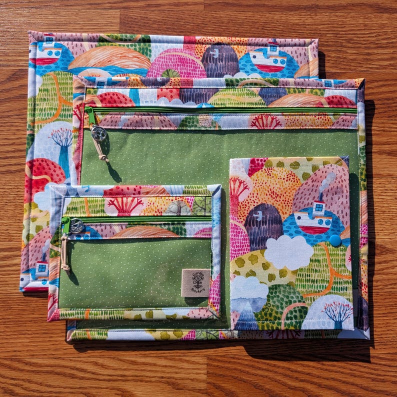 Puede incluir: Conjunto de tres bolsas de tela con un colorido estampado de paisaje con barcos, &aacute;rboles y colinas. Las bolsas tienen compartimentos verdes con cremallera y son de diferentes tama&ntilde;os. La bolsa m&aacute;s grande mide aproximadamente 30 cm x 23 cm.