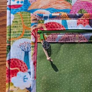 Puede incluir: Primer plano de una colorida bolsa de tela con cremallera verde y un dije de &aacute;rbol. La bolsa presenta un dise&ntilde;o de paisaje con elementos rojos, azules y verdes. El bolsillo interior es verde con lunares blancos.