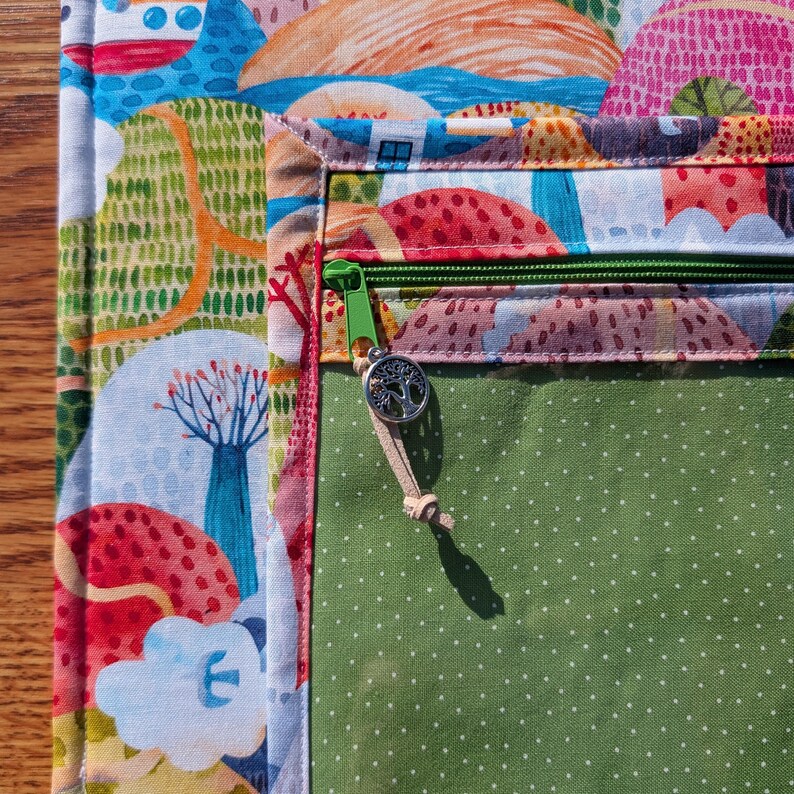 Puede incluir: Primer plano de una colorida bolsa de tela con cremallera verde y un dije de &aacute;rbol. La bolsa presenta un dise&ntilde;o de paisaje con elementos rojos, azules y verdes. El bolsillo interior es verde con lunares blancos.