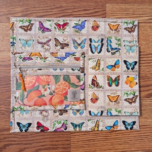 May include: A quilted fabric organizer with a butterfly stamp pattern and a zippered pocket featuring orange slices. The organizer has a beige background with a grid of colorful butterfly illustrations.