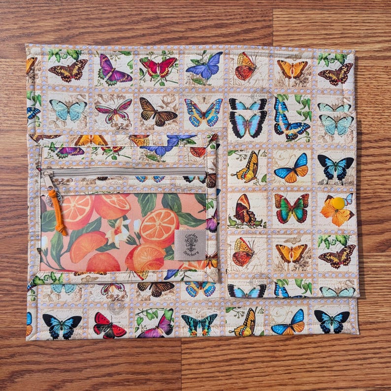 May include: A quilted fabric organizer with a butterfly stamp pattern and a zippered pocket featuring orange slices. The organizer has a beige background with a grid of colorful butterfly illustrations.