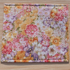 Puede incluir: Un mantel individual cuadrado con estampado floral que presenta un patr&oacute;n denso de varias flores en tonos rosa, amarillo, naranja y morado. Los bordes est&aacute;n cuidadosamente acabados con un borde cosido, creando un art&iacute;culo decorativo y funcional.