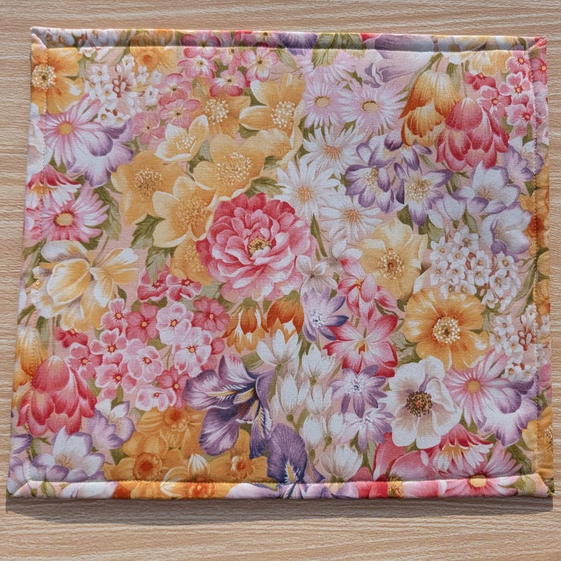 Puede incluir: Un mantel individual cuadrado con estampado floral que presenta un patr&oacute;n denso de varias flores en tonos rosa, amarillo, naranja y morado. Los bordes est&aacute;n cuidadosamente acabados con un borde cosido, creando un art&iacute;culo decorativo y funcional.