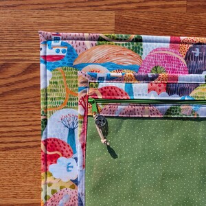 Puede incluir: Una colorida bolsa de tela acolchada con un dise&ntilde;o de paisaje caprichoso. La bolsa presenta una cremallera verde y un interior verde con lunares blancos. El exterior muestra un patr&oacute;n vibrante de casas, &aacute;rboles y elementos acu&aacute;ticos.