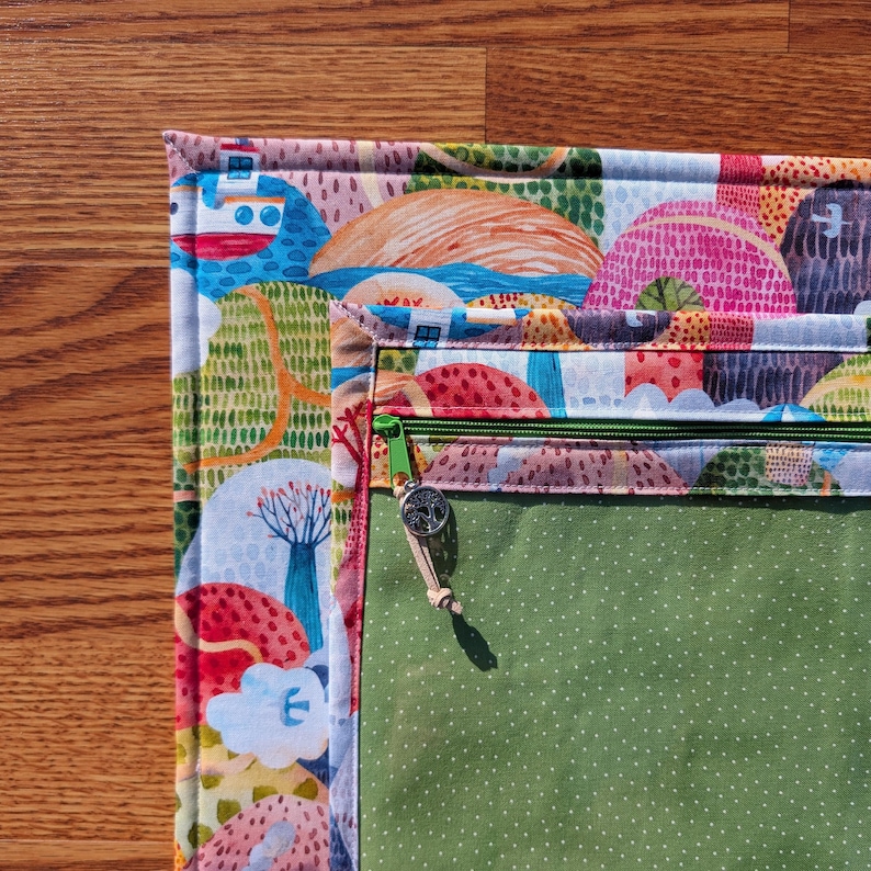 Puede incluir: Una colorida bolsa de tela acolchada con un dise&ntilde;o de paisaje caprichoso. La bolsa presenta una cremallera verde y un interior verde con lunares blancos. El exterior muestra un patr&oacute;n vibrante de casas, &aacute;rboles y elementos acu&aacute;ticos.