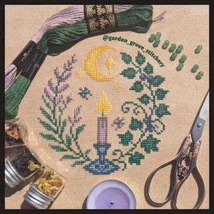 May include: A cross-stitch design depicting a crescent moon, stars, and a candle within a wreath of leaves and flowers. Embroidery floss, scissors, glass jars, and beads surround the design. The text "@garden_grove_stitchery" is visible.