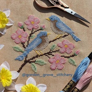 May include: Cross-stitch embroidery featuring two bluebirds perched on a flowering branch with pink blossoms and green leaves. The artwork is set against a tan fabric background, with embroidery floss and scissors visible.