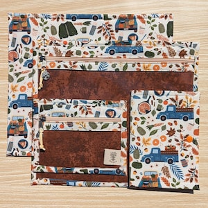 May include: A set of fall-themed zippered pouches in various sizes. The pouches feature a brown interior and a cream-colored exterior with a repeating pattern of blue trucks, pumpkins, and autumn leaves. The zippers are beige.