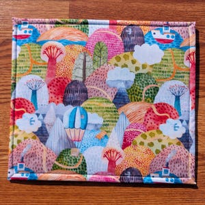 Puede incluir: Un art&iacute;culo de tela rectangular con un dise&ntilde;o de paisaje caprichoso. El patr&oacute;n presenta colinas, &aacute;rboles, nubes, un globo aerost&aacute;tico y barcos en tonos de rosa, verde, azul y naranja. El art&iacute;culo tiene un borde fino cosido.