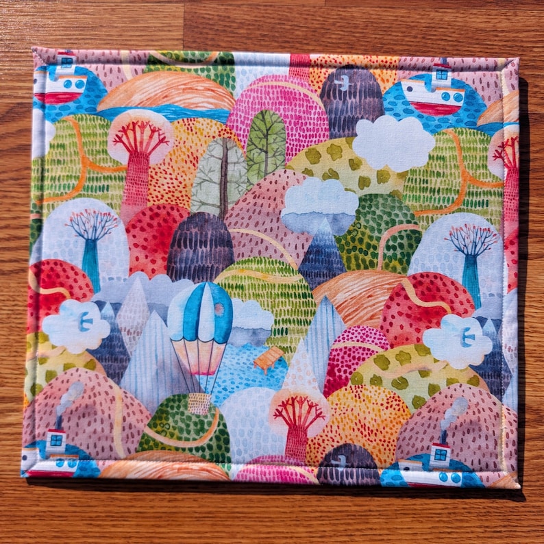 Puede incluir: Un art&iacute;culo de tela rectangular con un dise&ntilde;o de paisaje caprichoso. El patr&oacute;n presenta colinas, &aacute;rboles, nubes, un globo aerost&aacute;tico y barcos en tonos de rosa, verde, azul y naranja. El art&iacute;culo tiene un borde fino cosido.