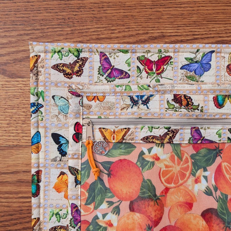 May include: A multi-pocket organizer with a butterfly and orange slice pattern. The top section features a cream-colored fabric with butterfly illustrations. The bottom section has a peach-colored fabric with orange slices. A zipper and orange leather pull are visible.