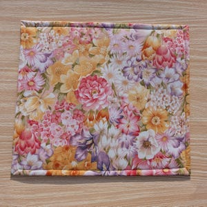 Puede incluir: Un mantel individual cuadrado con estampado floral que presenta una vibrante variedad de flores en tonos rosa, amarillo, naranja y morado. Los bordes est&aacute;n cuidadosamente acabados con un borde a juego, creando una pieza decorativa y funcional para la mesa.