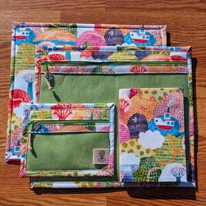 Puede incluir: Un conjunto de tres bolsas de tela coloridas con un patr&oacute;n de paisaje caprichoso. Cada bolsa tiene una cremallera verde y un bolsillo transparente. La bolsa m&aacute;s grande es rectangular, la del medio cuadrada y la m&aacute;s peque&ntilde;a rectangular.