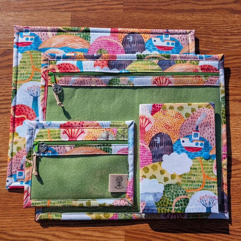 Puede incluir: Un conjunto de tres bolsas de tela coloridas con un patr&oacute;n de paisaje caprichoso. Cada bolsa tiene una cremallera verde y un bolsillo transparente. La bolsa m&aacute;s grande es rectangular, la del medio cuadrada y la m&aacute;s peque&ntilde;a rectangular.