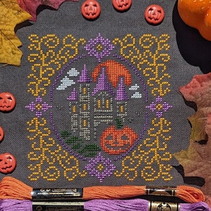 May include: Cross-stitch design depicting a Halloween castle with a pumpkin and full moon. The design is framed by a decorative border in gold and purple. Orange and purple embroidery floss and pumpkin-shaped buttons surround the design.