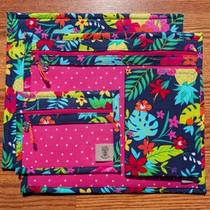May include: Set of four zippered pouches with a tropical floral pattern. The pouches feature a navy blue background with colorful flowers, leaves, and pineapples. The interior is pink with white polka dots. Each pouch has a pink zipper and a small tag.